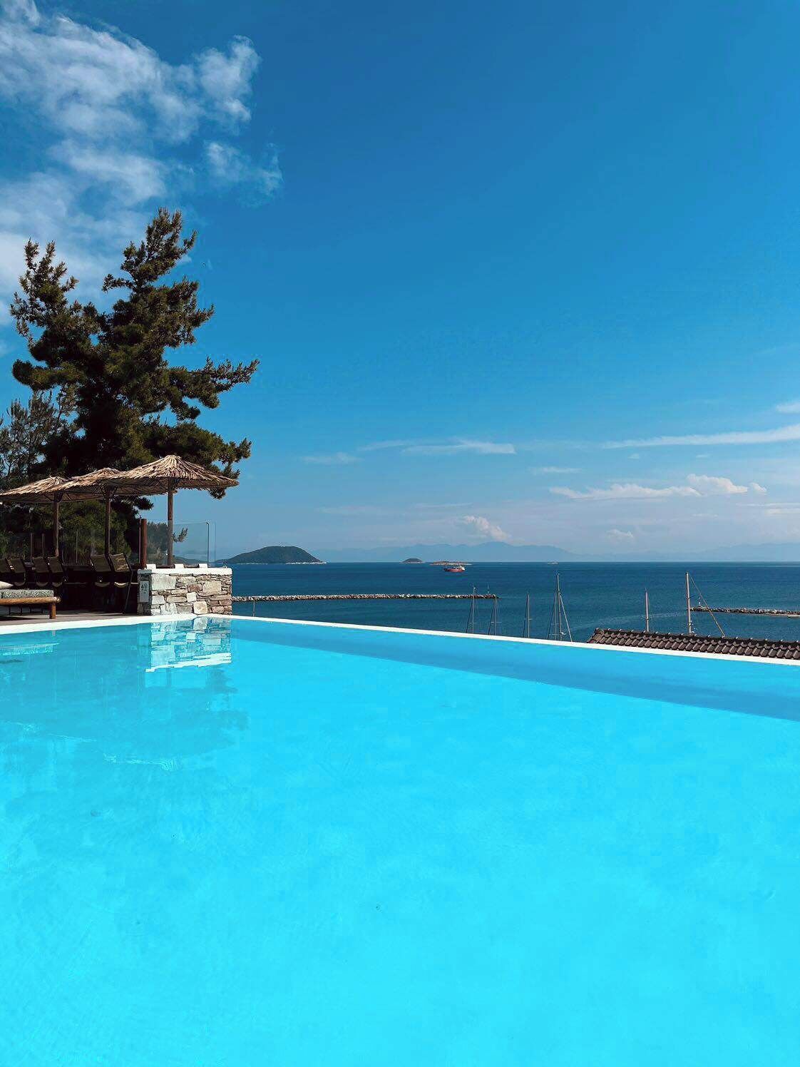 A clear blue pool with a stone structure and lounge chairs on one side, overlooking a calm sea and distant islands.