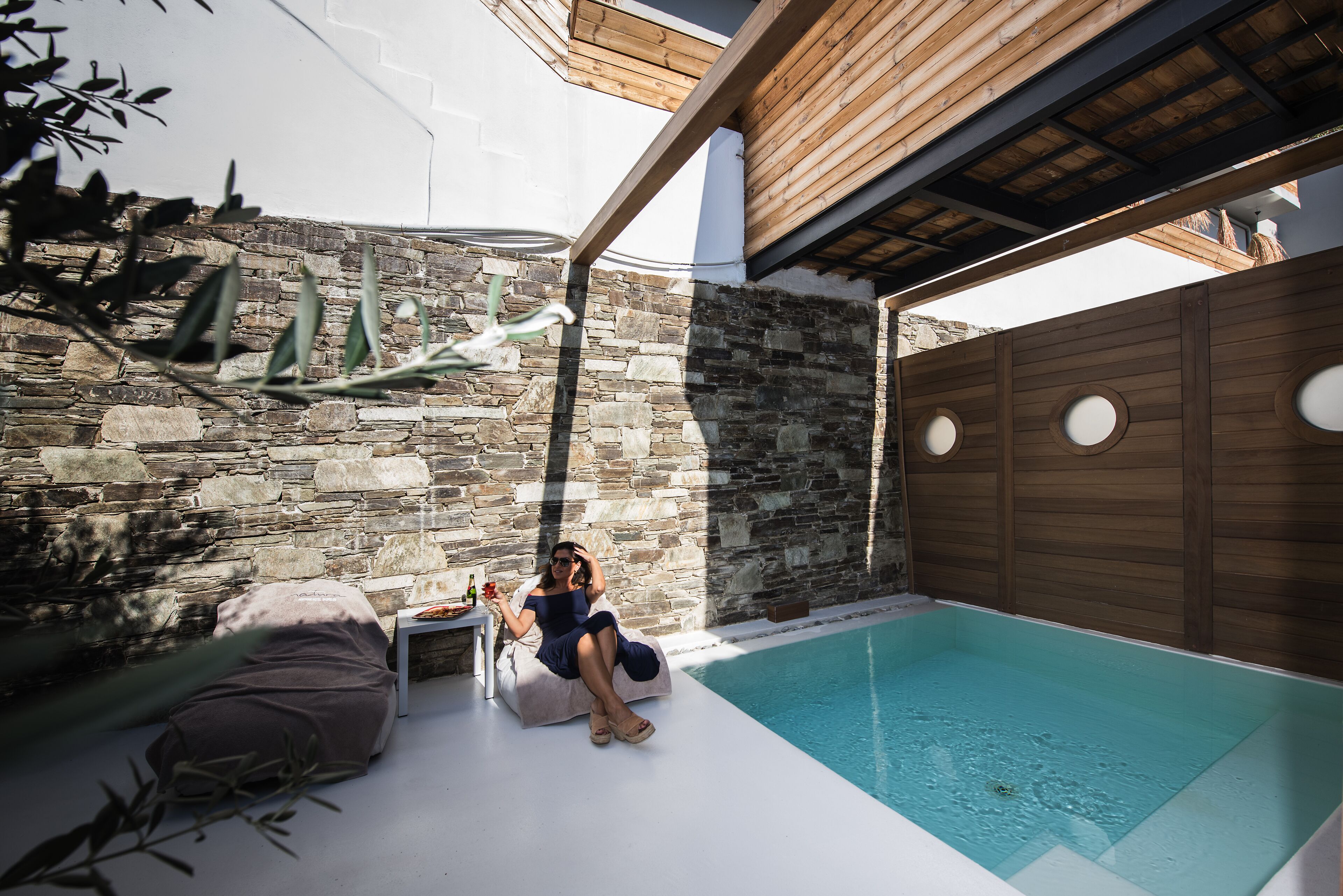 Una persona relajándose junto a una piscina con un muro de piedra y paneles de madera en un entorno exterior moderno.