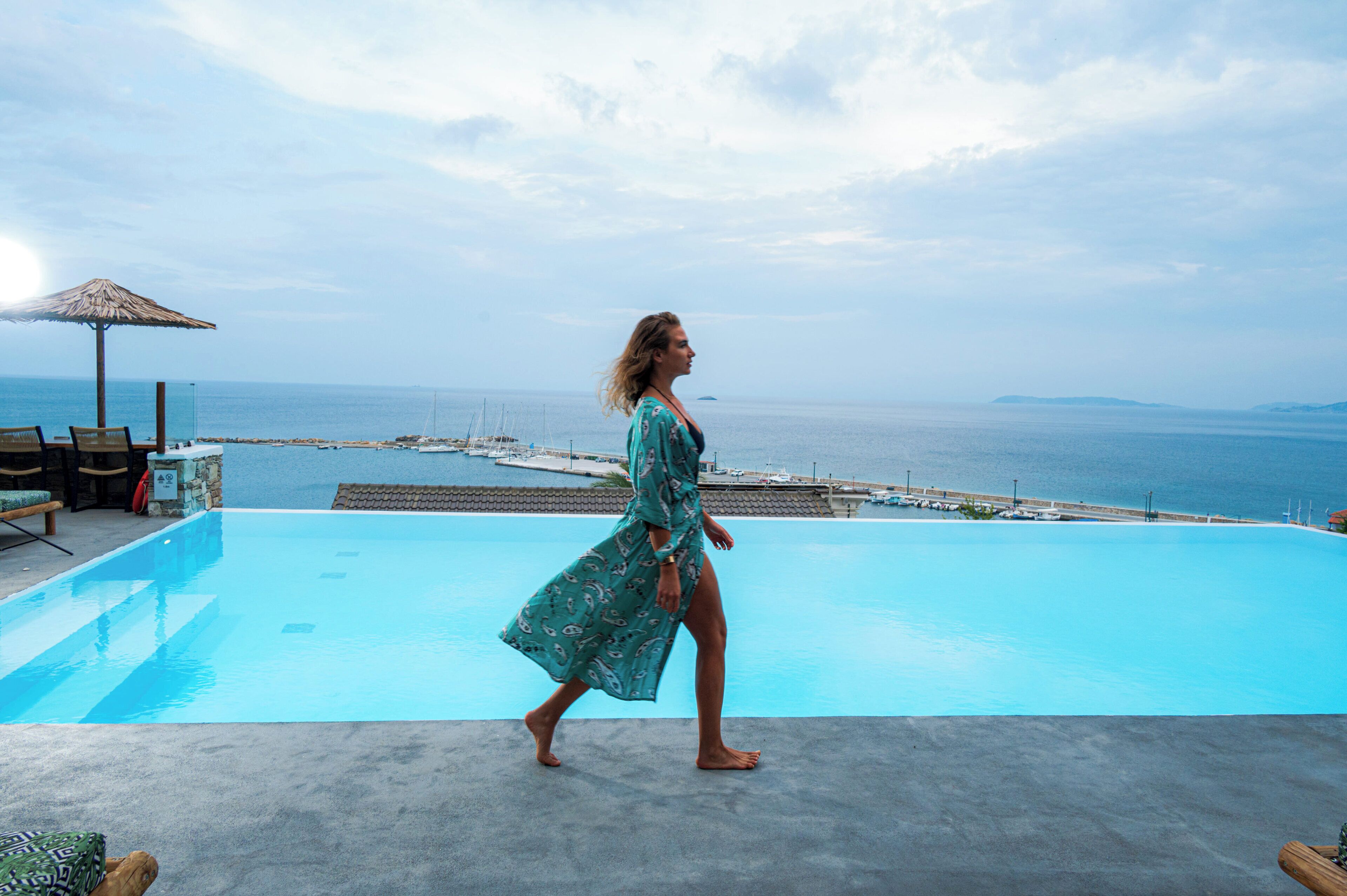 Eine Frau geht am Pool entlang mit Blick auf das Meer und einen Steg im Hintergrund.