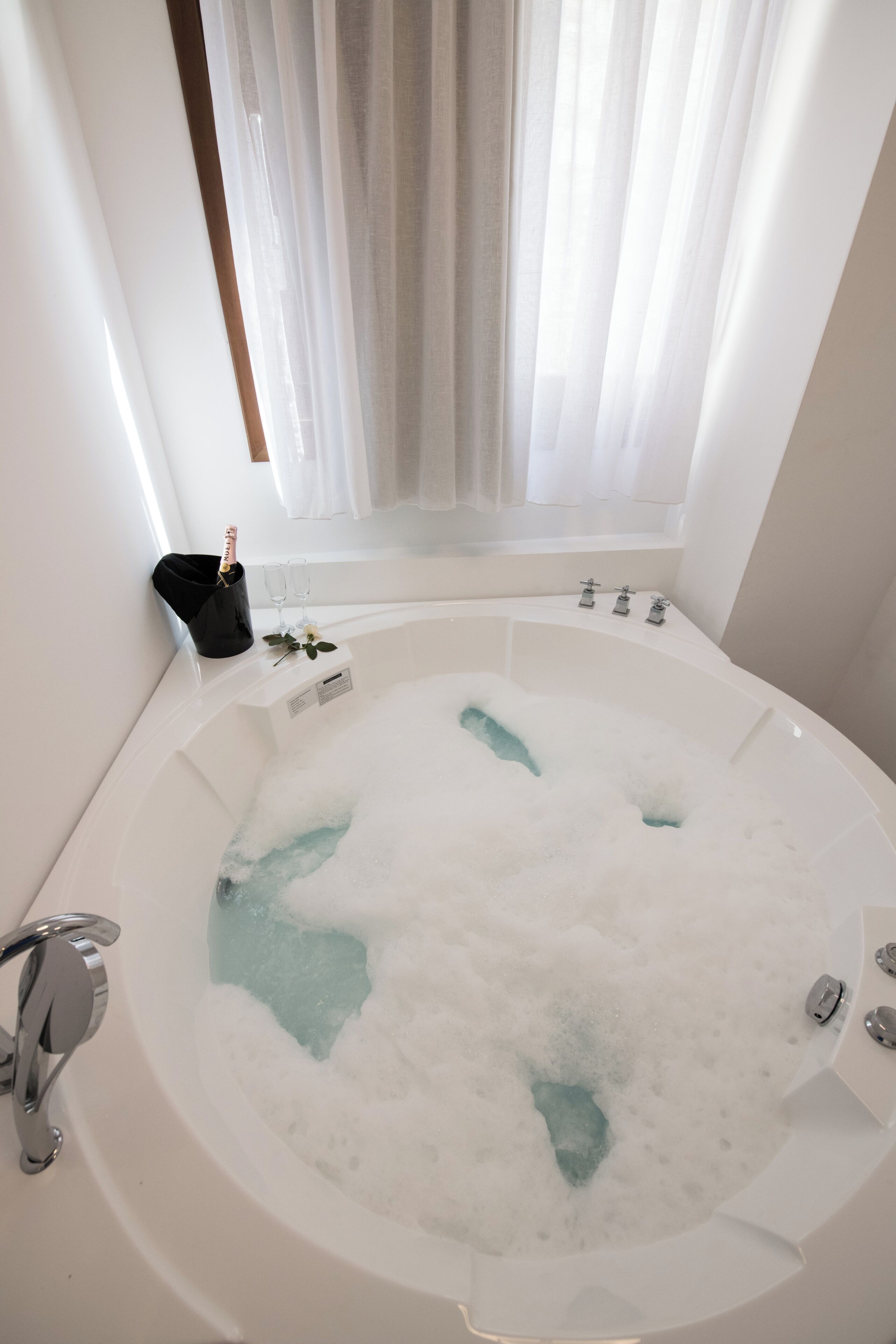A large, white bathtub filled with bubbles, a champagne bottle, and two glasses on a tray.