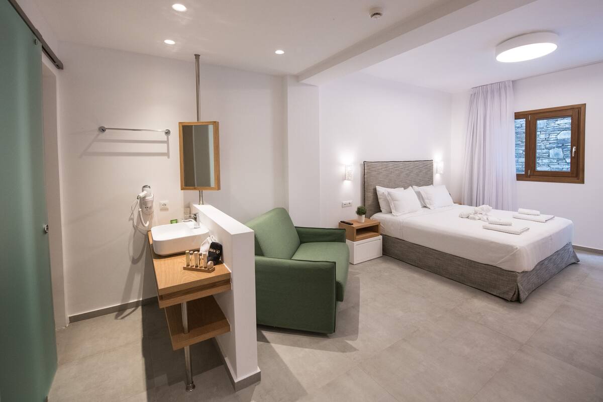Une chambre d’hôtel moderne avec un canapé vert, un lit avec du linge de maison blanc, une petite table de chevet, un miroir et une fenêtre avec un cadre en pierre.
