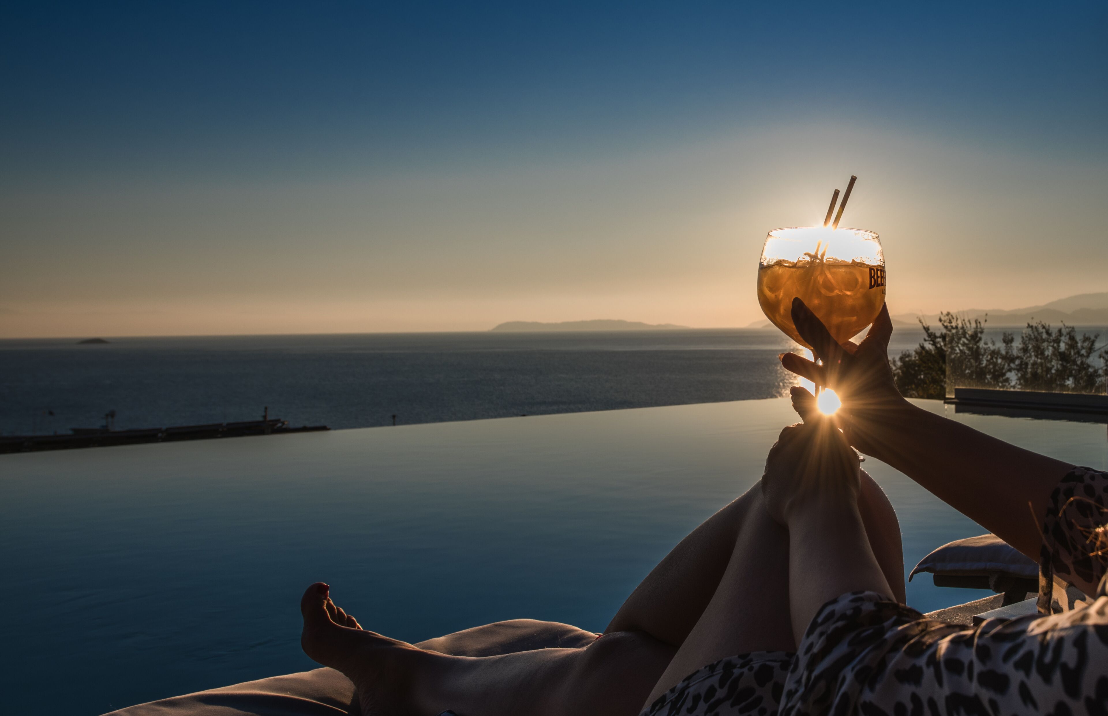 A person relaxing by a pool with a drink, overlooking a body of water and distant land.