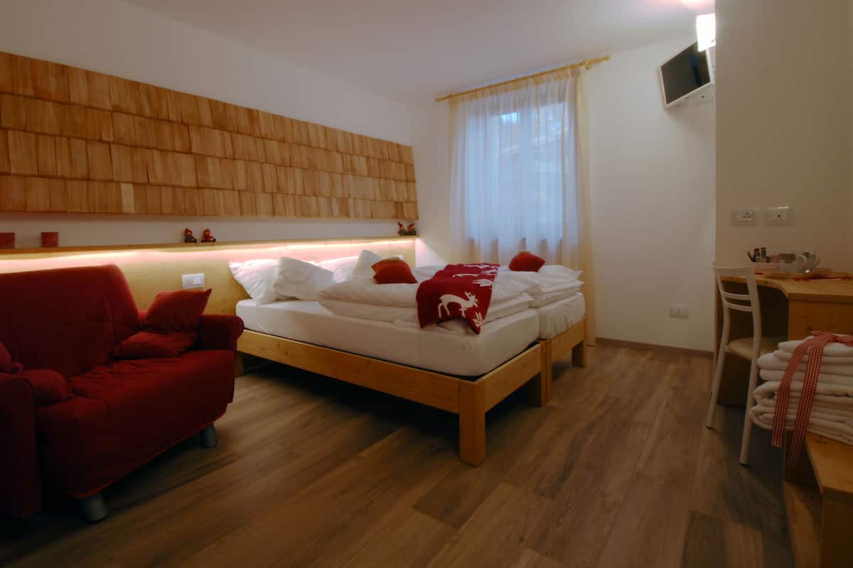 A bedroom with a wooden headboard, a bed with white bedding and red pillows, a red armchair, a wooden desk with a chair, and a window with sheer curtains.