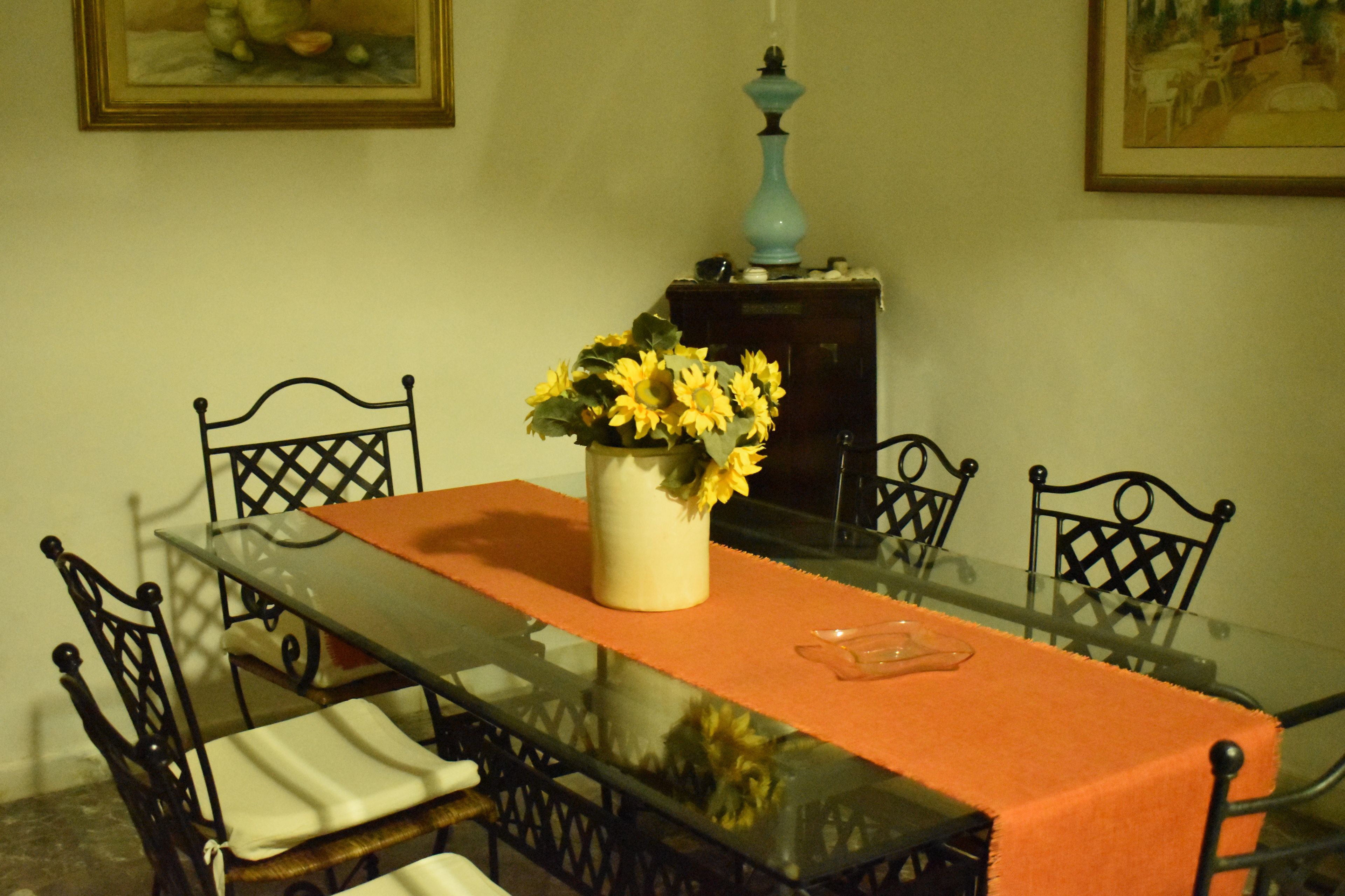 A dining table with a glass top and orange table runner, surrounded by black metal chairs.