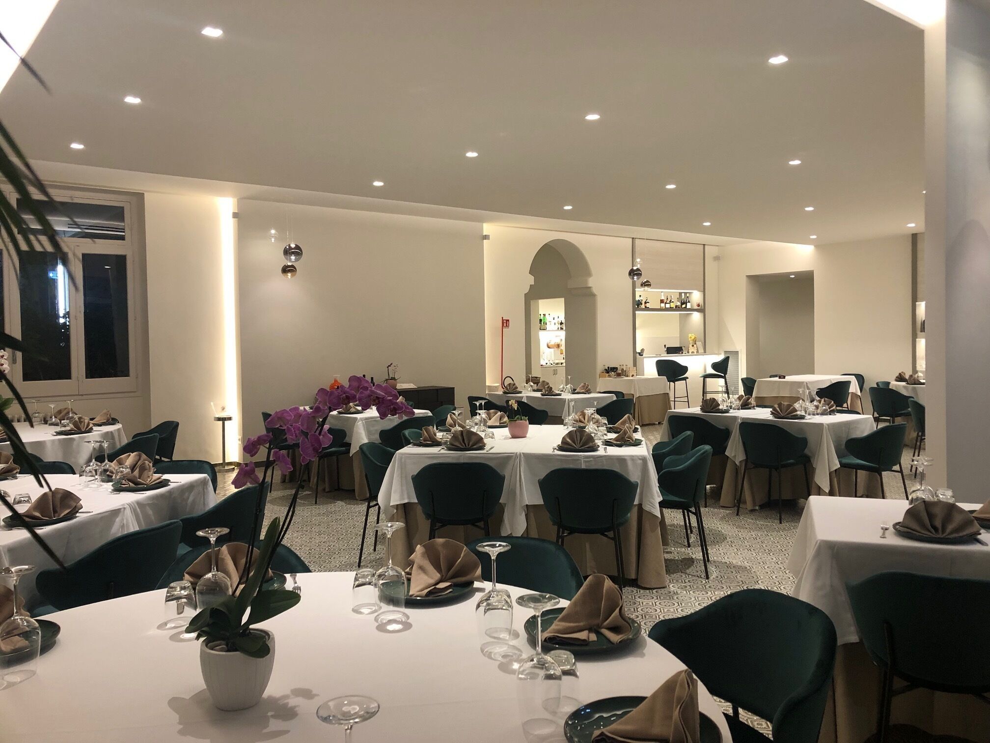 Un comedor con varias mesas dispuestas para invitados, con manteles blancos, sillas verdes y orquídeas moradas como decoración.