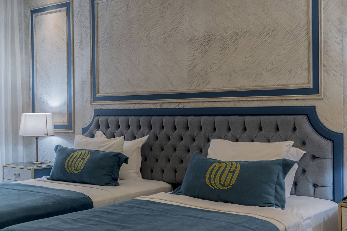 Une chambre d’hôtel avec une tête de lit capitonnée, deux lits avec une parure de lit bleue assortie et une table de chevet avec une lampe.