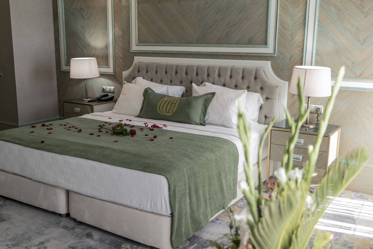 Un lit bien fait, avec une couvre-lit verte et des oreillers blancs, une tête de lit capitonnée et une table de chevet avec une lampe.