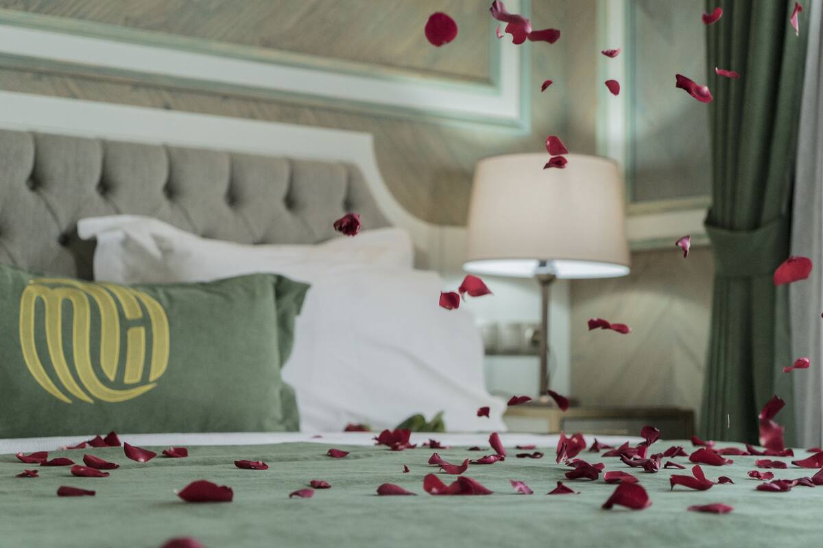 Un lit bien fait, avec un oreiller vert et une couverture blanche, une lampe de chevet et des pétales de rose éparpillés sur le lit.