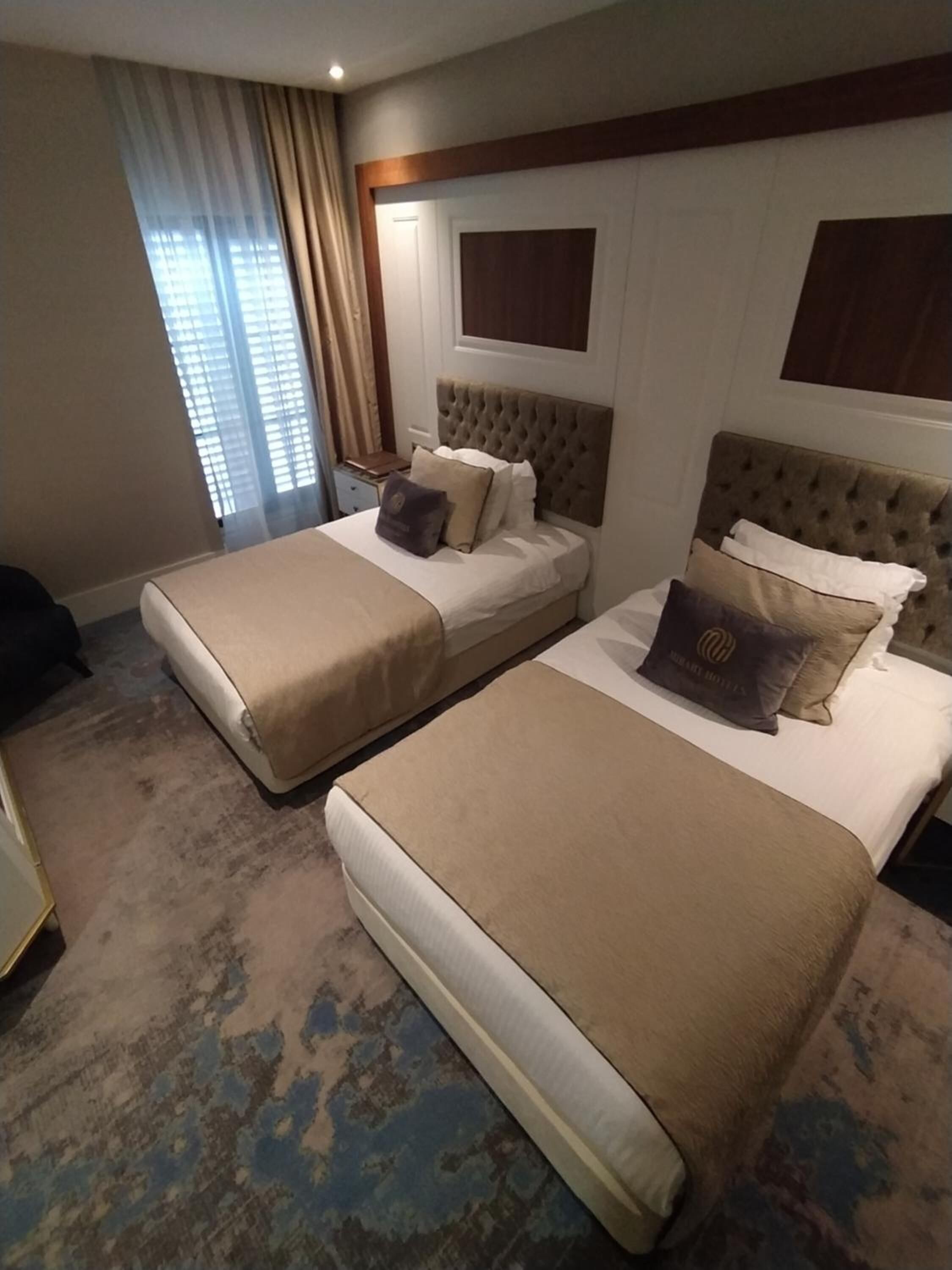 A hotel room with two beds, a large window with curtains, a wooden headboard, and a patterned carpet.