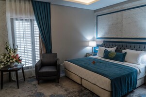 Habitación de hotel con una cama grande, una silla, una mesita de noche y un ventanal con cortinas.