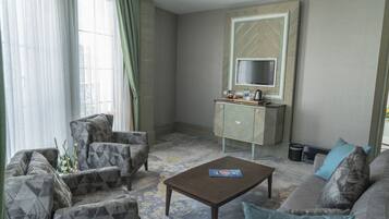 Una sala de estar con un sofá, un sillón y una mesa de centro. Hay un televisor sobre un mueble bajo, y una ventana con cortinas.