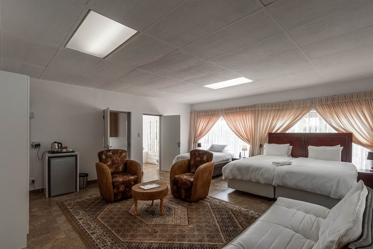 Habitación de hotel con cama, dos sillones, una mesita, un escritorio con lámpara, un refrigerador minibar y un ventanal grande con cortinas.