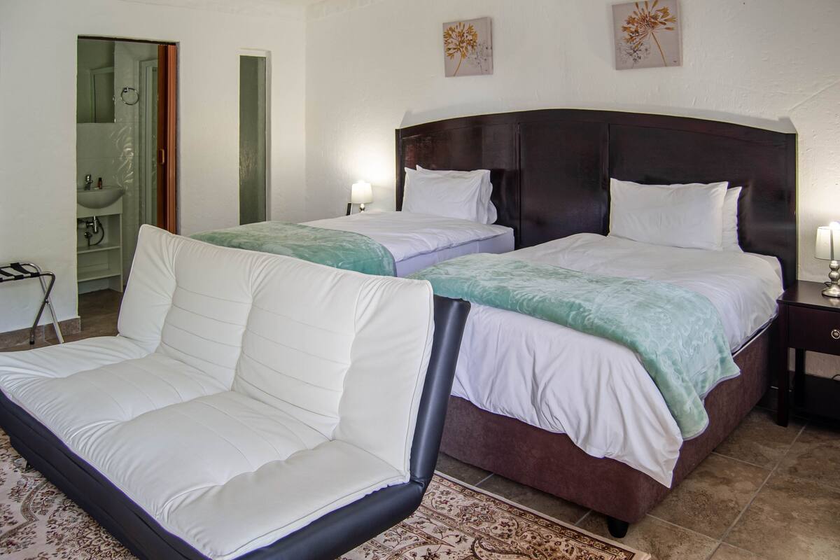 Una habitación de hotel con dos camas, un sofá, un baño con lavamanos y espejo, y obras de arte en las paredes.