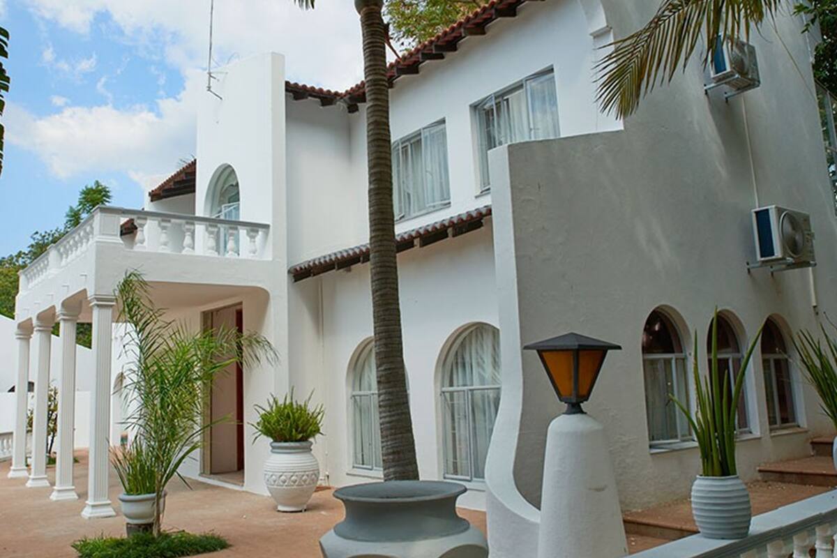 Un edificio blanco con ventanas arqueadas y un balcón, rodeado de plantas en macetas y un cielo despejado.
