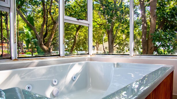 A glass-enclosed hot tub with a wooden bench, surrounded by large windows offering a view of trees and a garden. - Graceland Conference & Lifestyle Centre (Akasia)