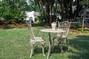 Un jardín con una mesa redonda y dos sillas ornamentales, rodeado de abundante follaje y un edificio blanco al fondo.