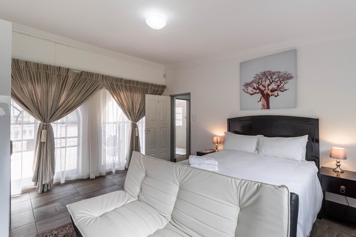 Una habitación de hotel con una cama grande, una mesita de noche con lámpara, una ventana con cortinas y un cuadro enmarcado en la pared.