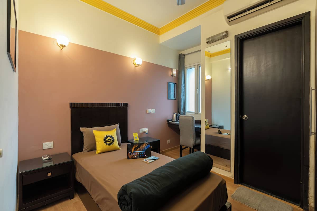A hotel room with a bed, bedside table, a desk with a chair, a door, and a mirror.