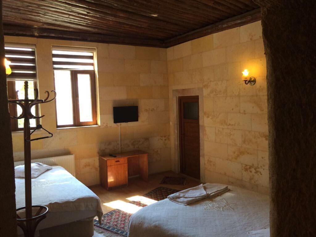 A room with two beds, a wooden ceiling, a stone wall, a small wooden desk, and a wall-mounted TV.