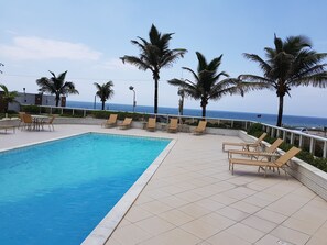 A swimming pool with lounge chairs and palm trees, overlooking the ocean. - Apartamento na praia de Armação. (Salvador)