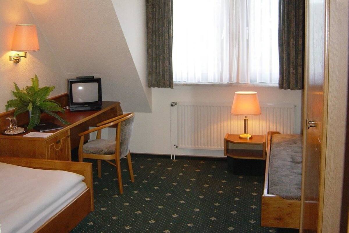 Habitación de hotel con cama, escritorio con computadora, silla, mesita de noche con lámpara y ventana con cortinas.