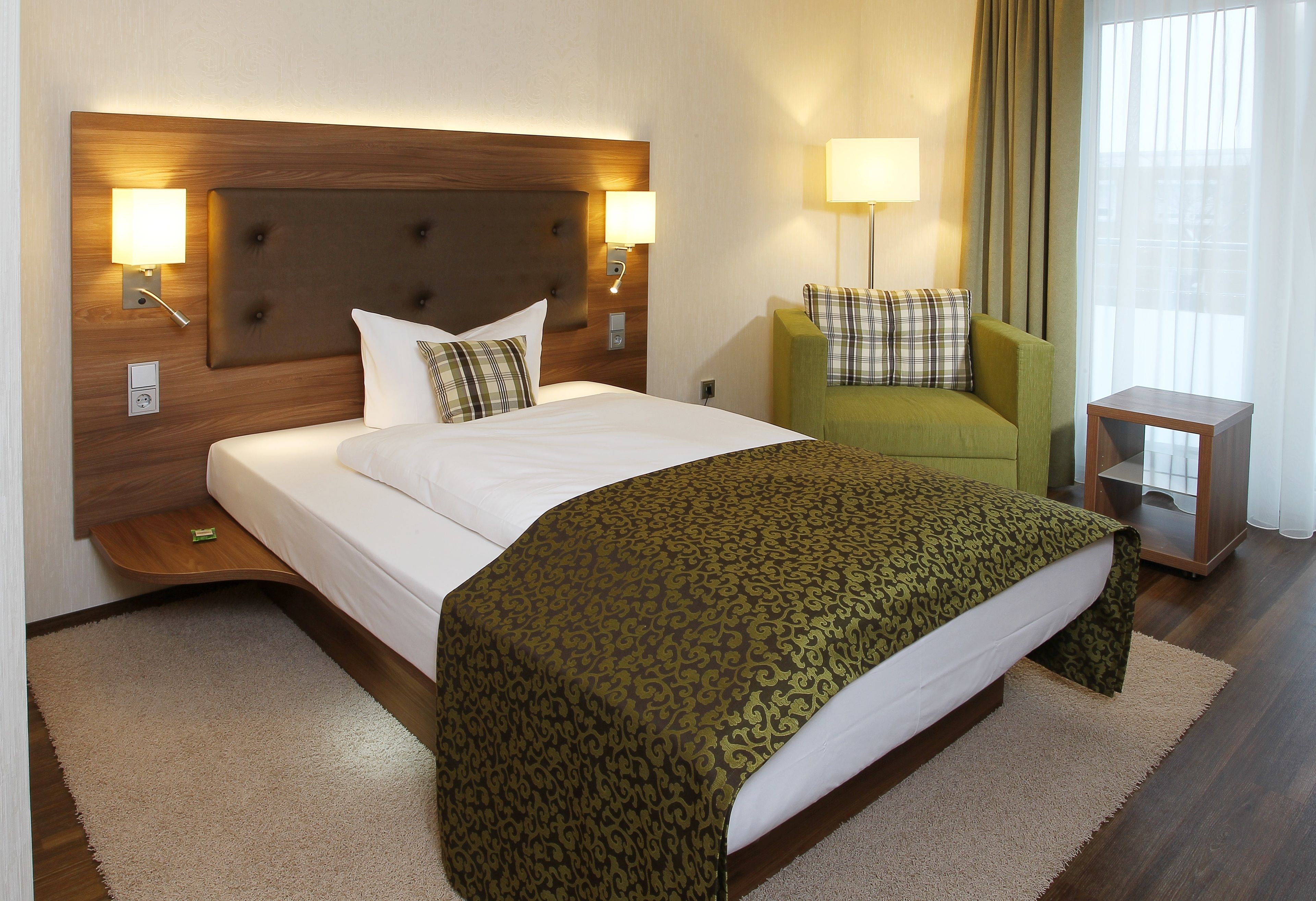 Una habitación de hotel con una cama grande, un sillón verde, una mesita de noche y una lámpara.