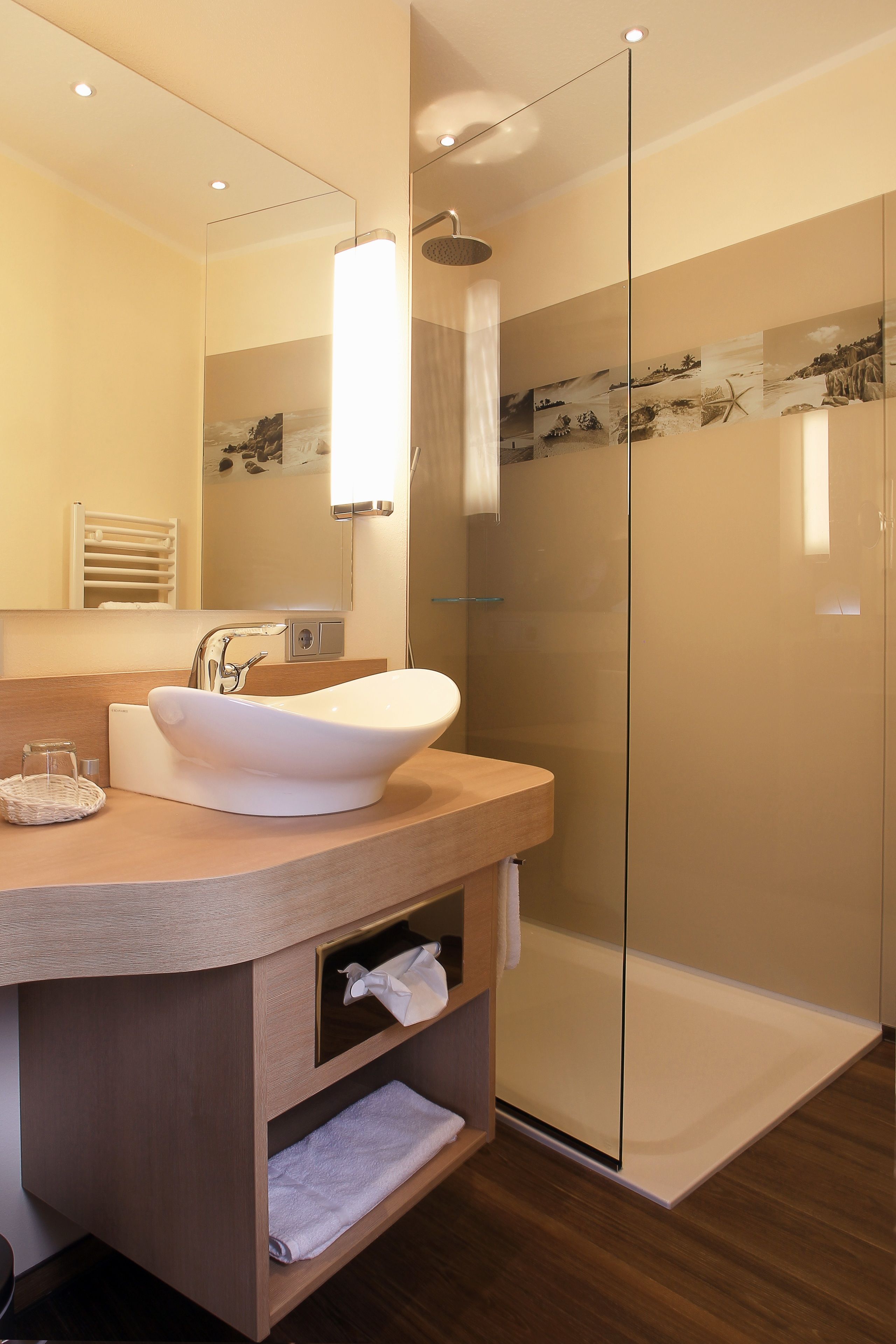 A modern bathroom with a large mirror, a white sink, a glass shower enclosure, and wooden flooring.
