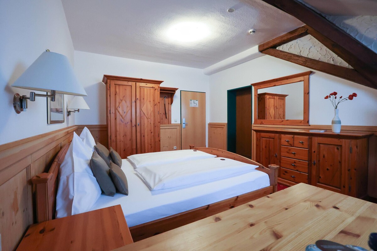 A wooden bed with white bedding, a wooden dresser, a mirror, and a lamp in a room with wooden walls and a sloped ceiling.