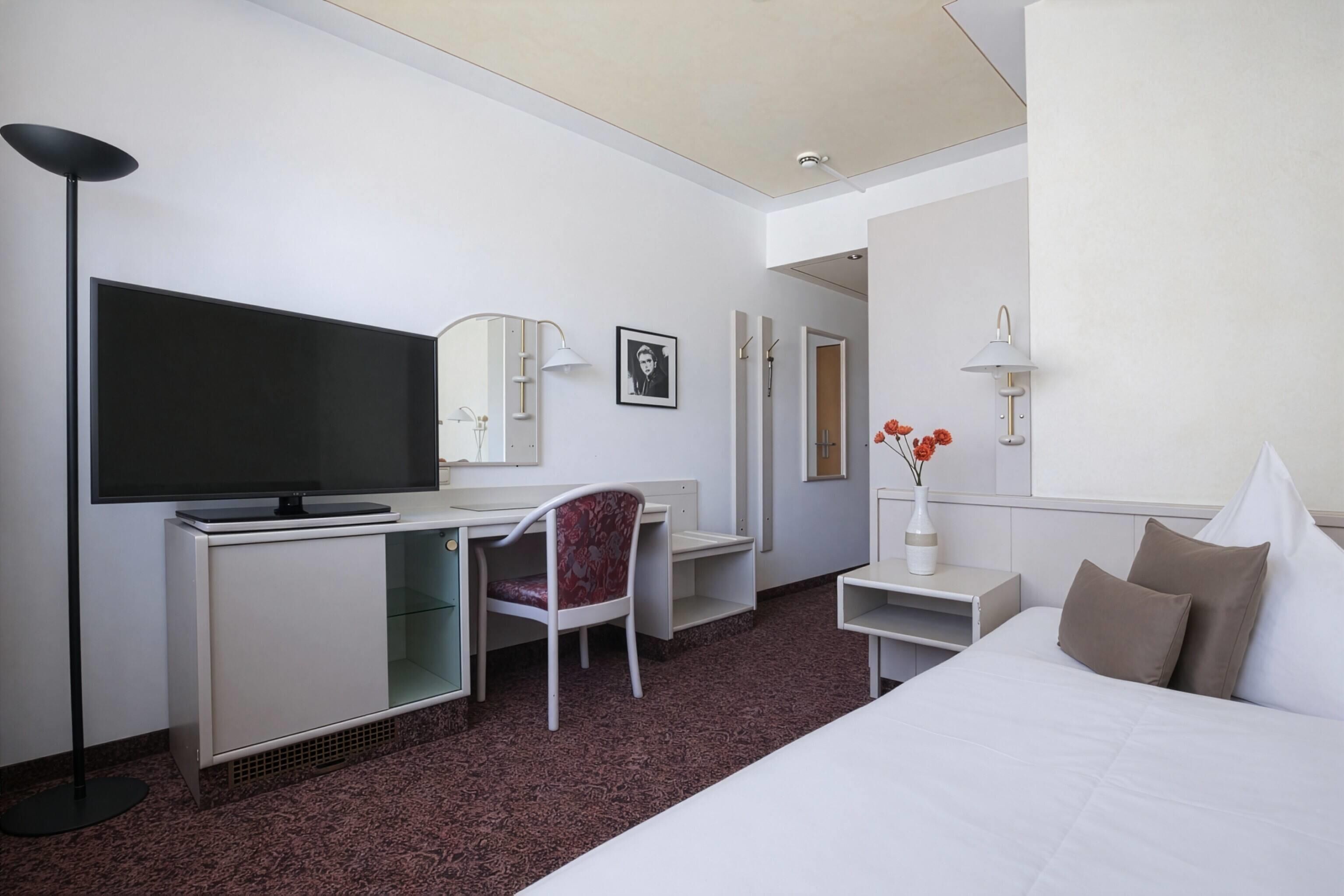 Une chambre d’hôtel comprenant un lit, un bureau, une télévision, une chaise, un miroir, une lampe et un vase avec des fleurs.