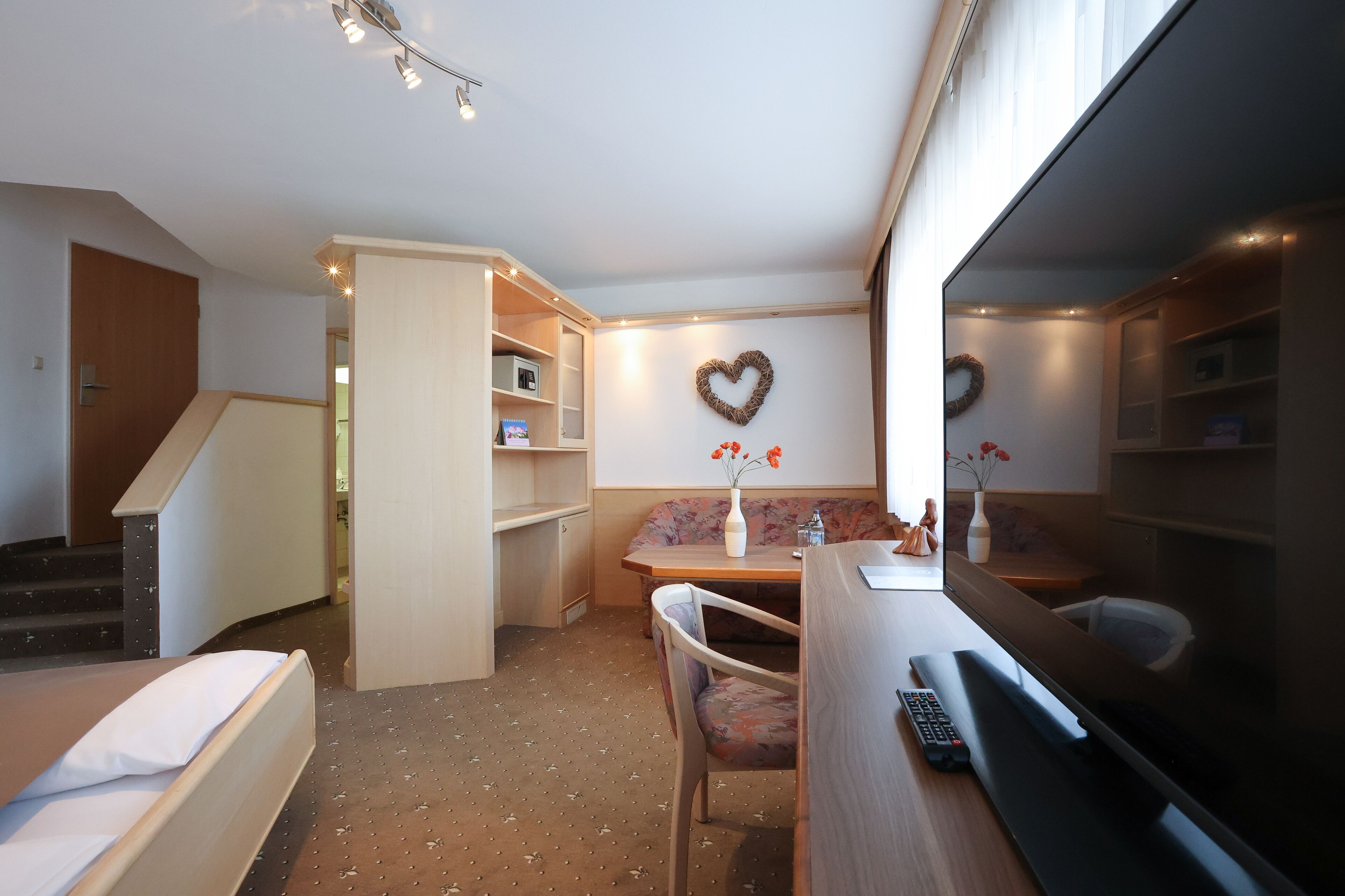 Een kleine kamer met een bed, een bureau met een computer, een stoel, een klein tafeltje met een vaas en een televisie.
