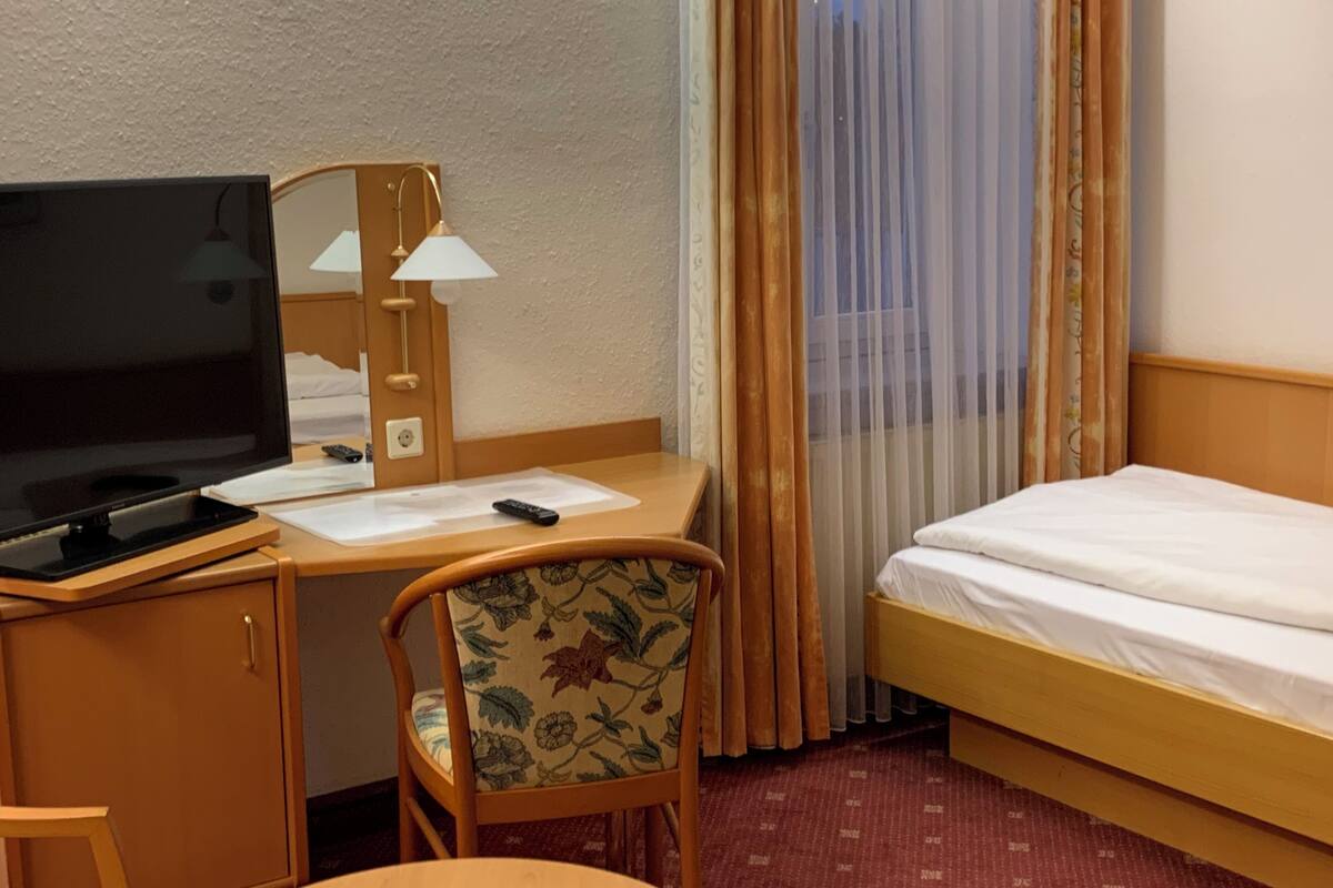 A hotel room with a bed, a desk with a TV, a chair, and a small table.