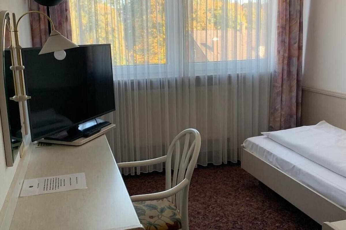 A hotel room with a bed, a desk with a TV, a chair, and a window with curtains.