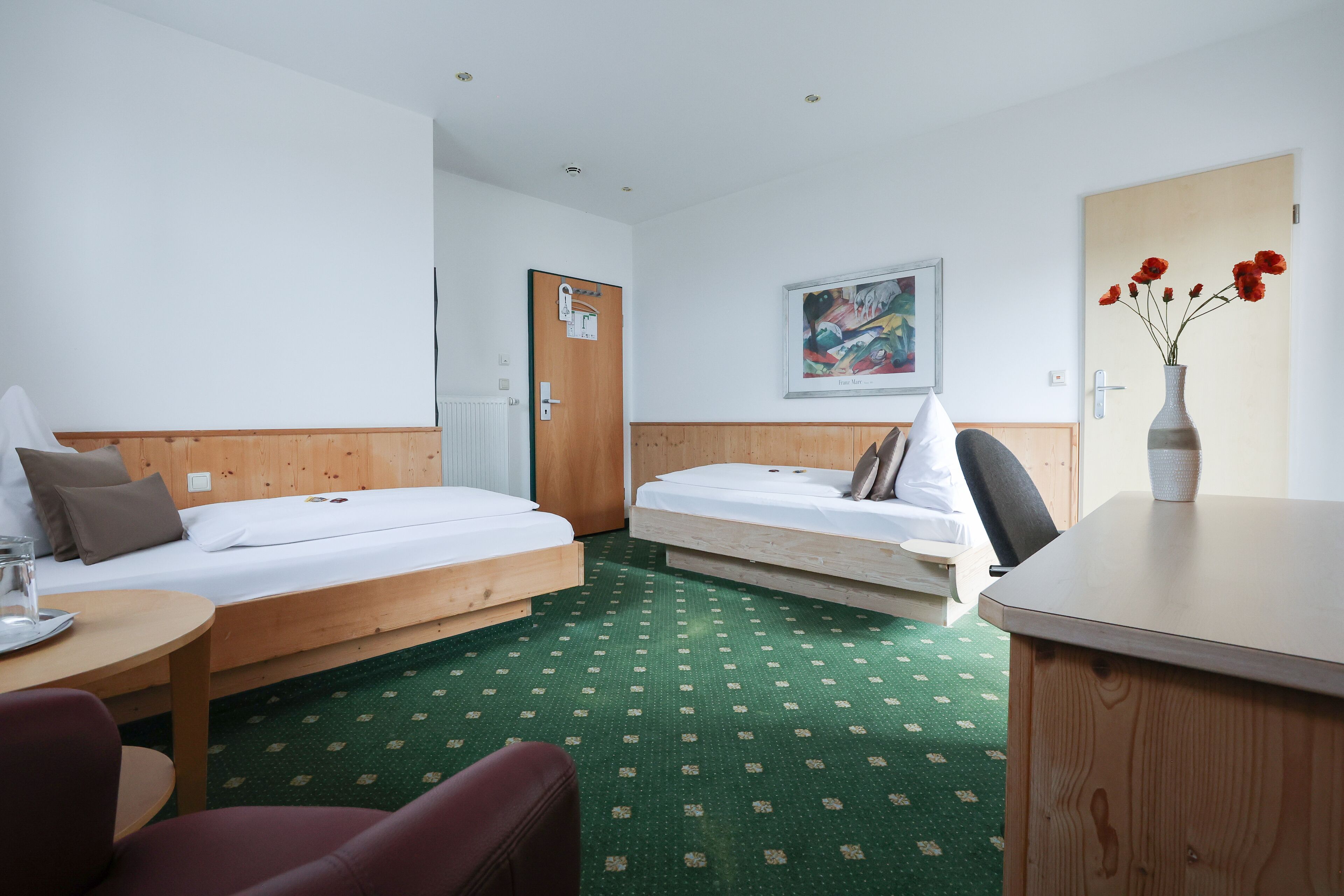 A hotel room with two beds, a desk, a chair, a radiator, a door, and a painting on the wall.