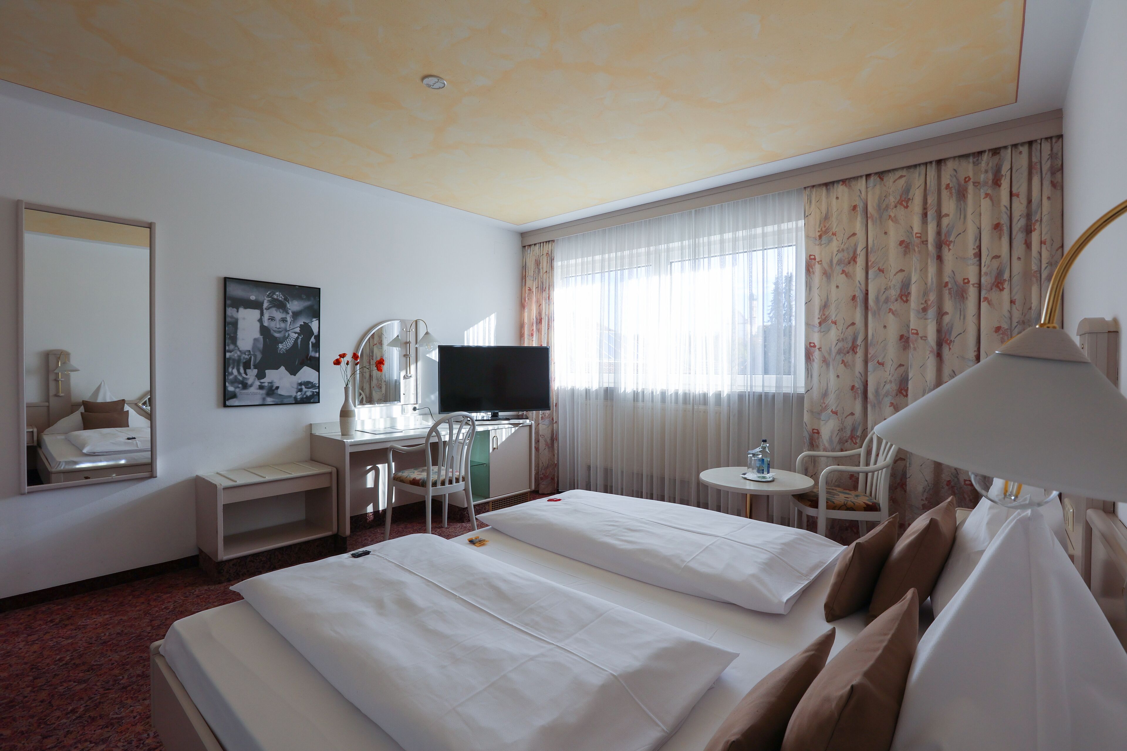 Habitación de hotel con dos camas, un escritorio con televisor, una silla, una mesita y una ventana con cortinas.