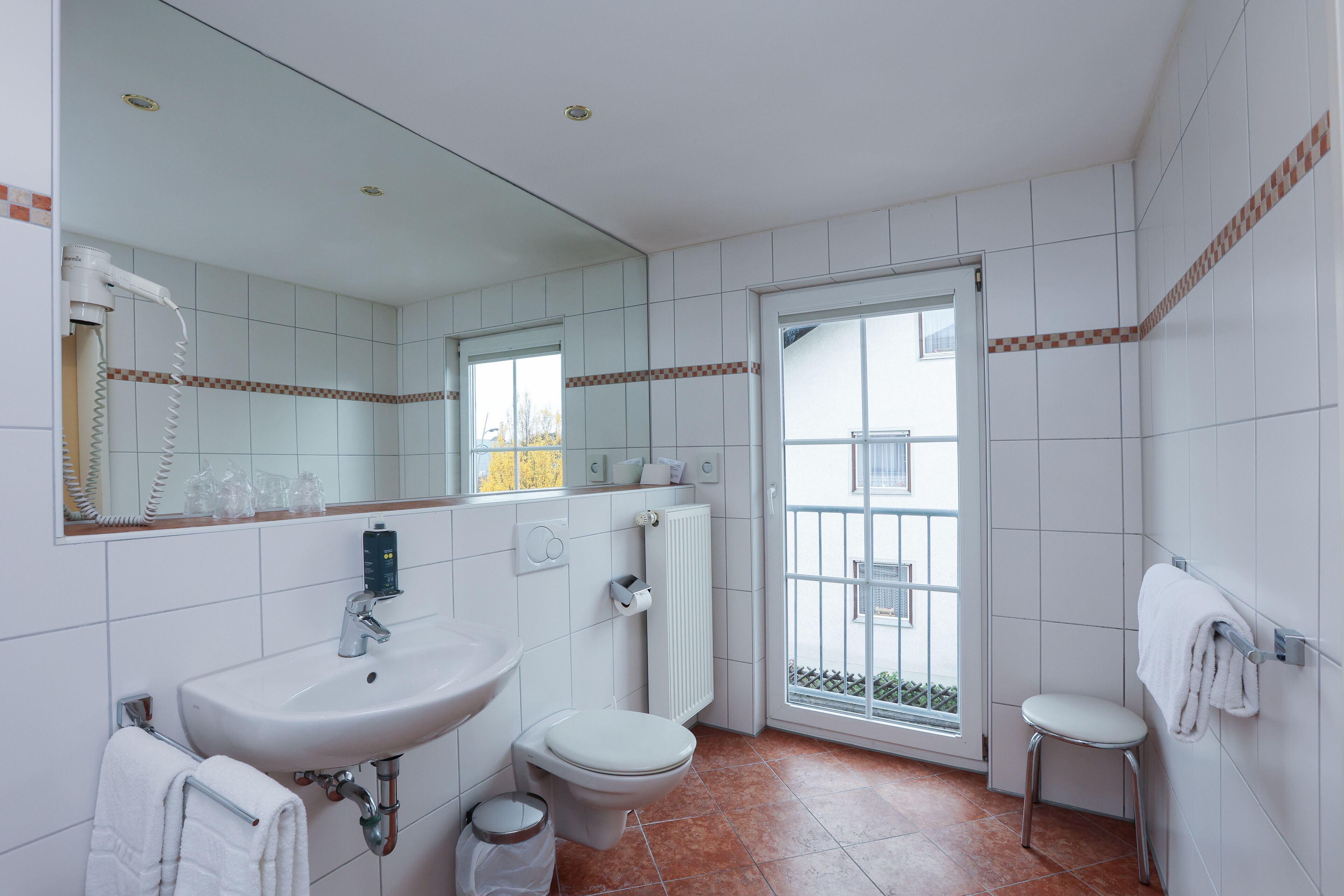 A bathroom with a sink, toilet, and a mirror. There is a towel rack and a trash bin.