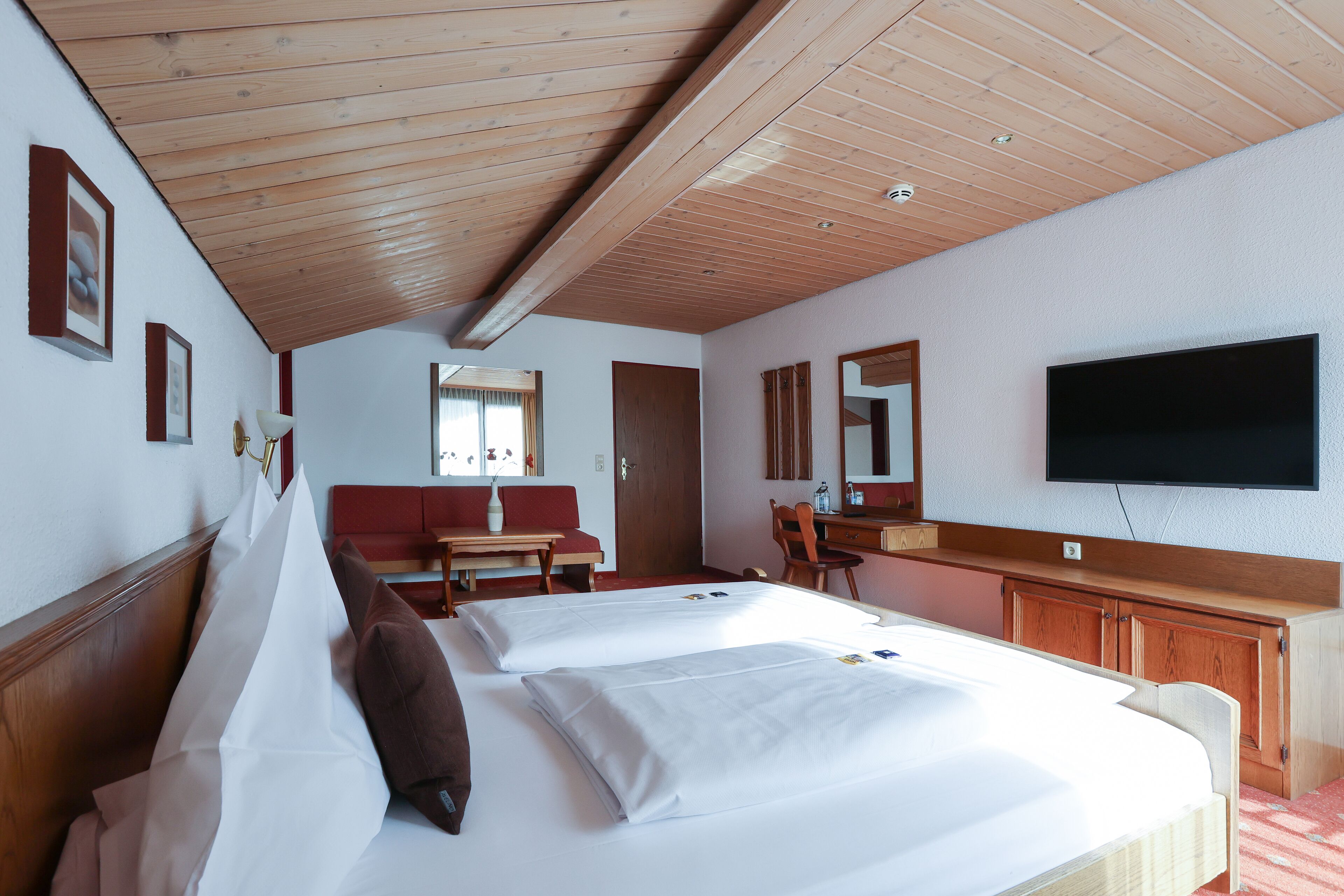 A hotel room with two beds, wooden ceiling, a TV, and a desk.