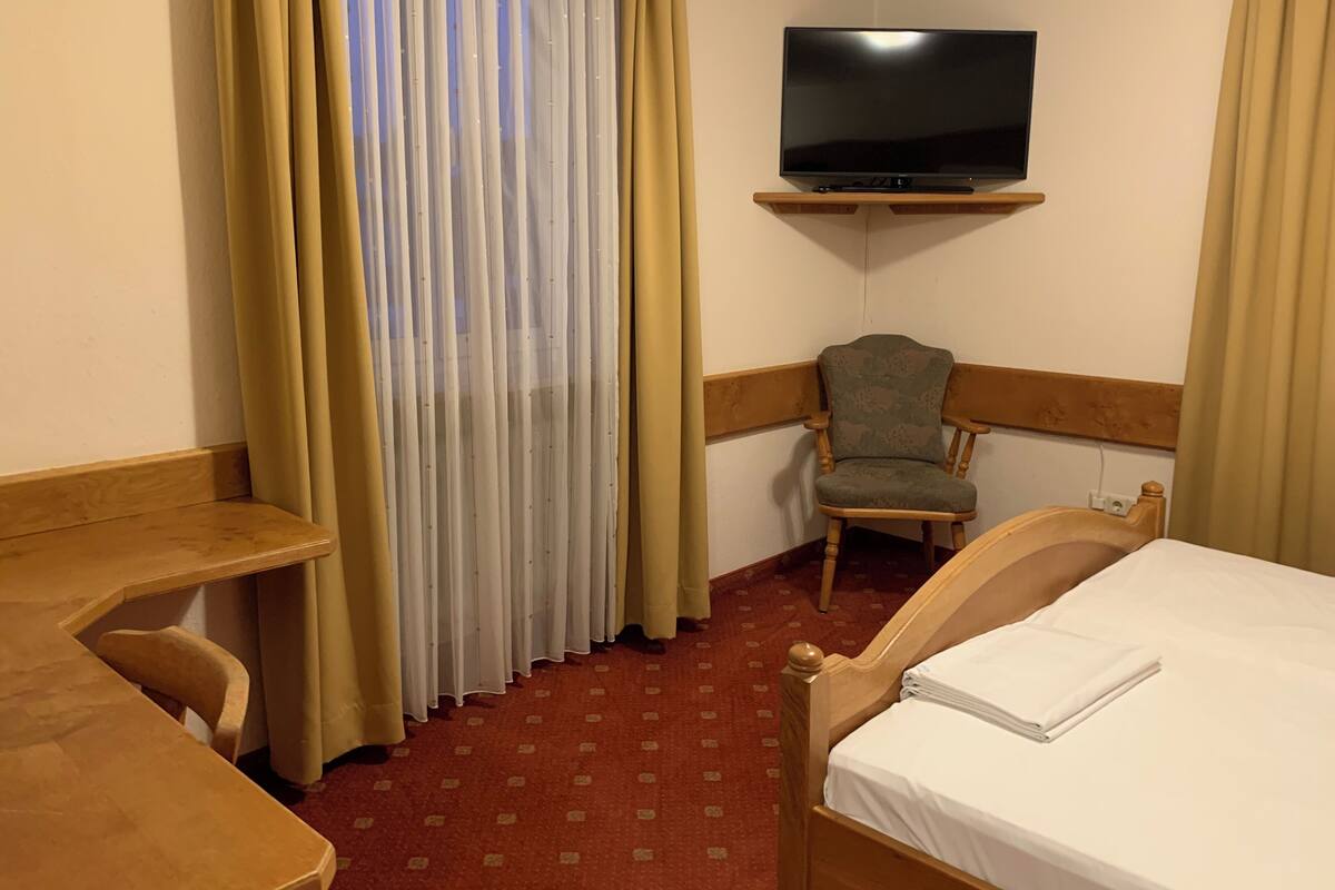 A hotel room with a bed, a chair, a desk, a TV mounted on the wall, and a window with curtains.