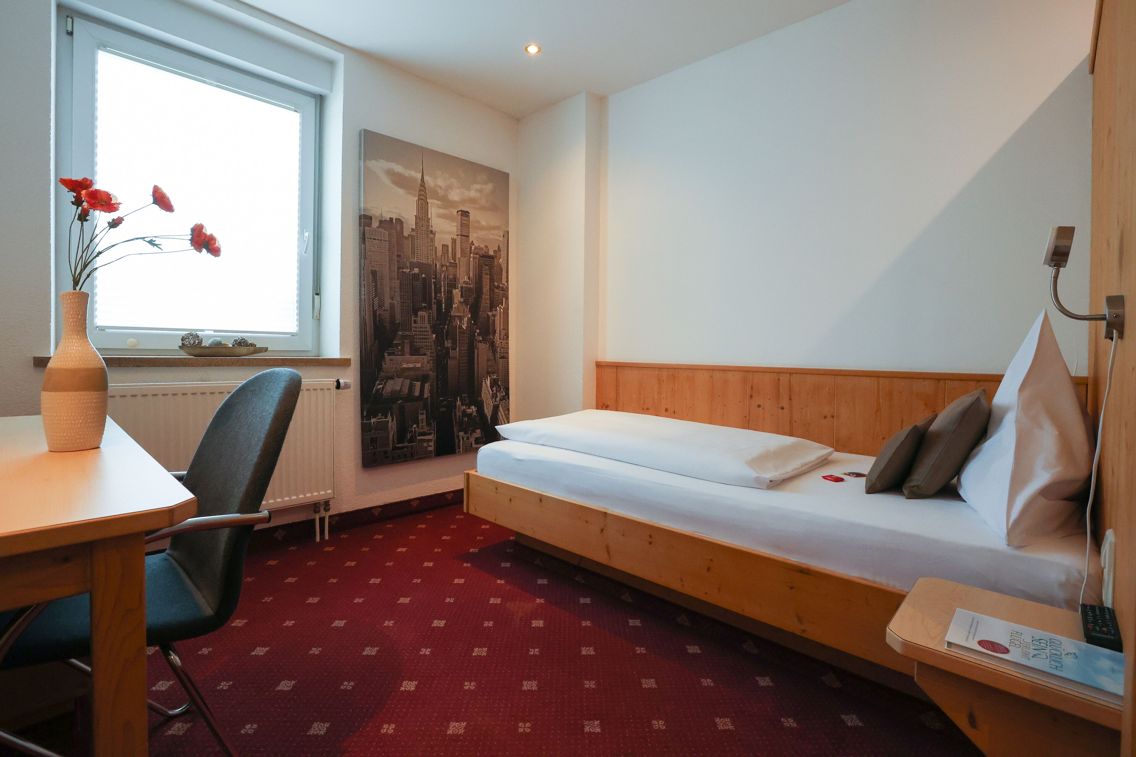 A hotel room with a wooden bed, a desk with a vase of flowers, a chair, and a cityscape mural on the wall.