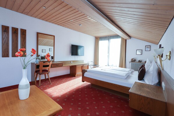 Habitación de hotel con techo de madera, una cama, un escritorio, una silla, un televisor, un espejo y un florero con flores.