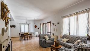 Een ruime woonkamer met betegelde vloer, een eethoek met tafel en stoelen, en een zithoek.