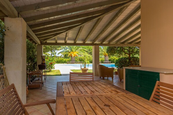 A covered outdoor patio with wooden furniture, a pool, and lush greenery in the background. - Villa Arenella con Piscina by Wonderful Italy (Siracusa)