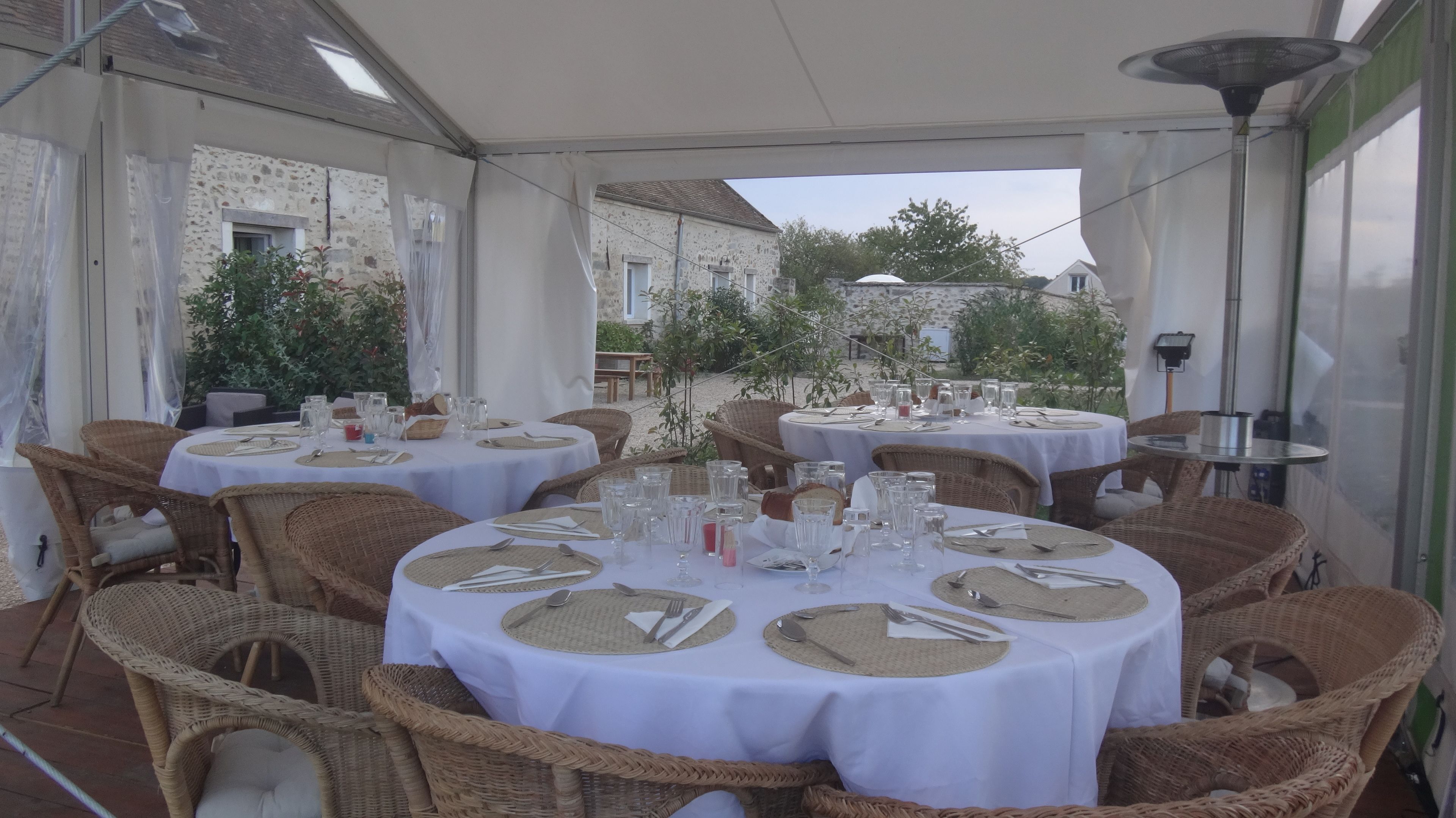 Un área de comedor al aire libre con mesas redondas cubiertas con manteles blancos, sillas de mimbre y cristalería.