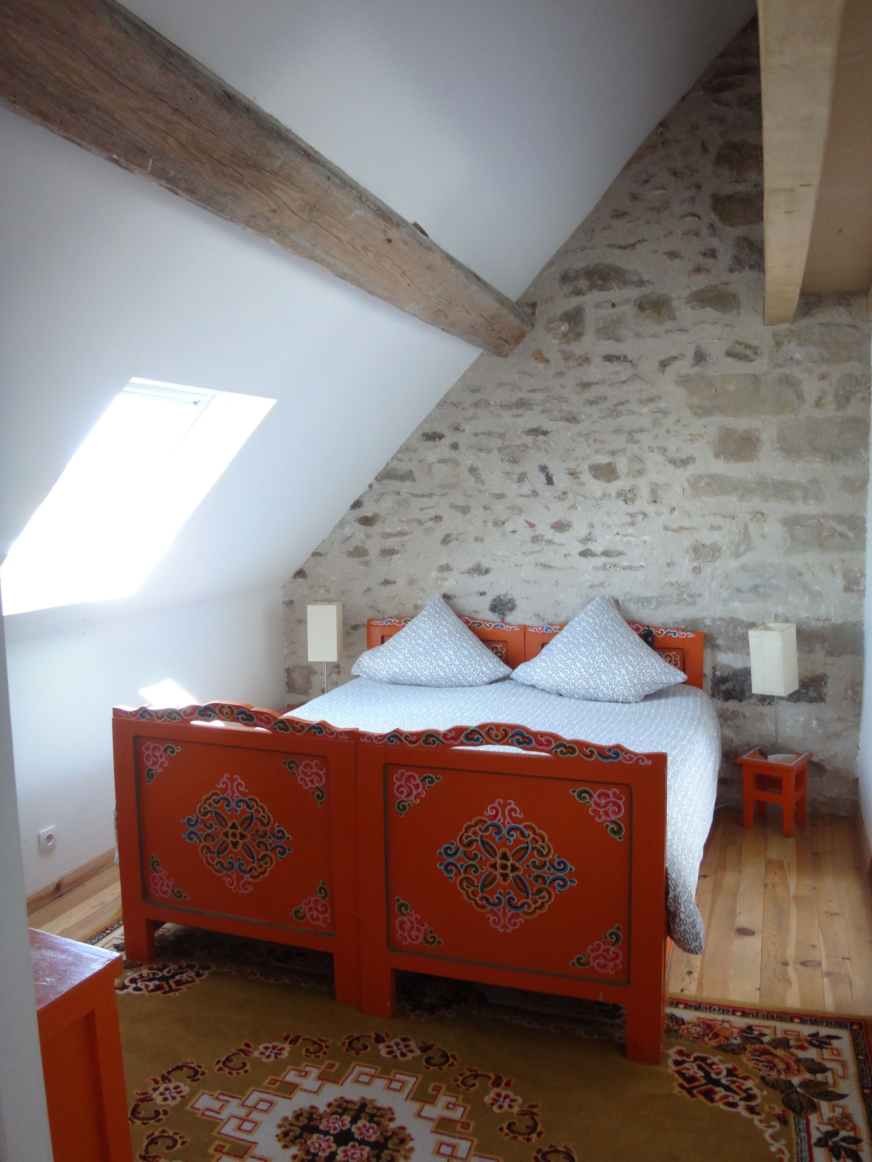 Un dormitorio con techo inclinado, pared de piedra, estructura de cama de madera y alfombra estampada.