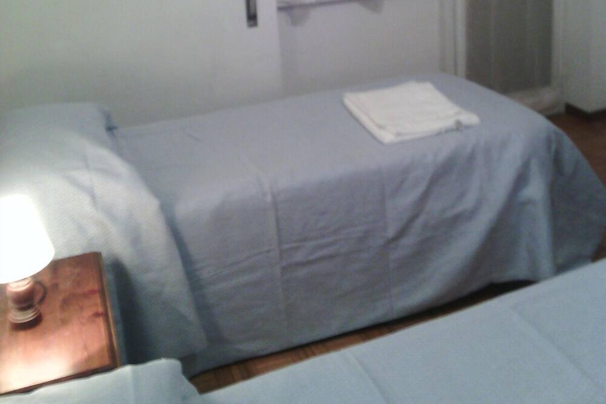 Ein Einzelbett mit weißer Decke, ein Nachttisch mit Lampe und ein Fenster mit Vorhängen.