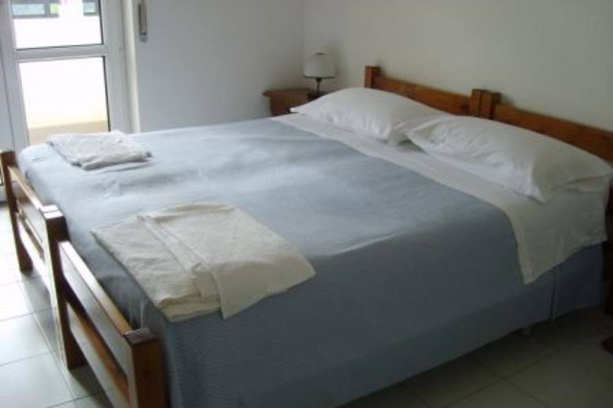 Ein ordentlich bezogenes Bett mit blauer Bettdecke und weißen Kissen, ein Holz-Kopfschild und ein Nachttischchen mit einer Lampe.