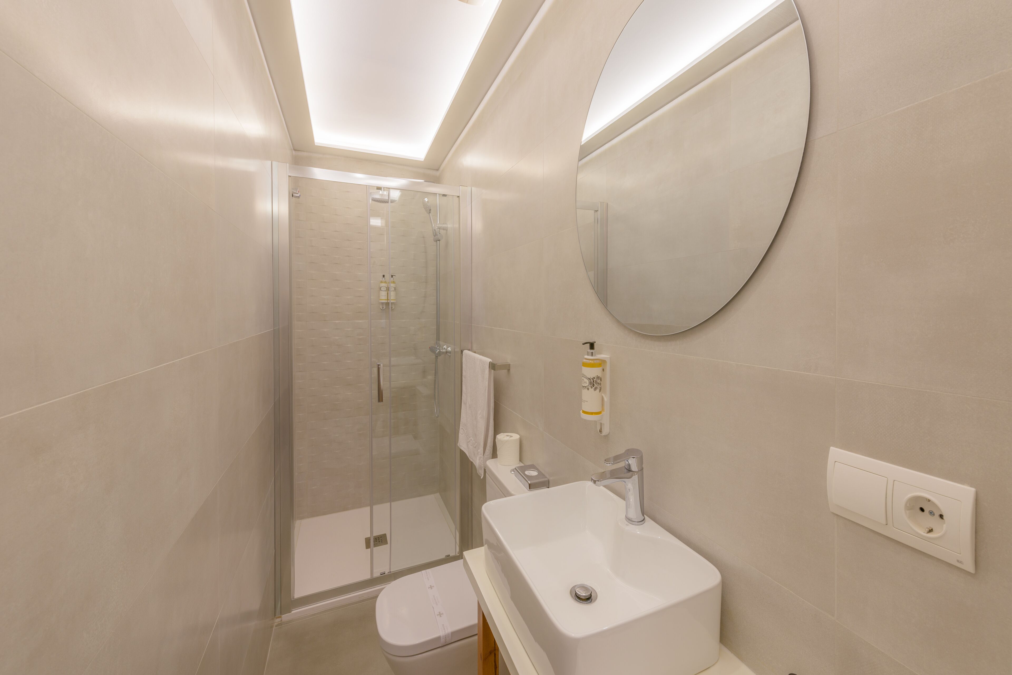 A modern bathroom with a glass shower enclosure, a white sink, and a round mirror.