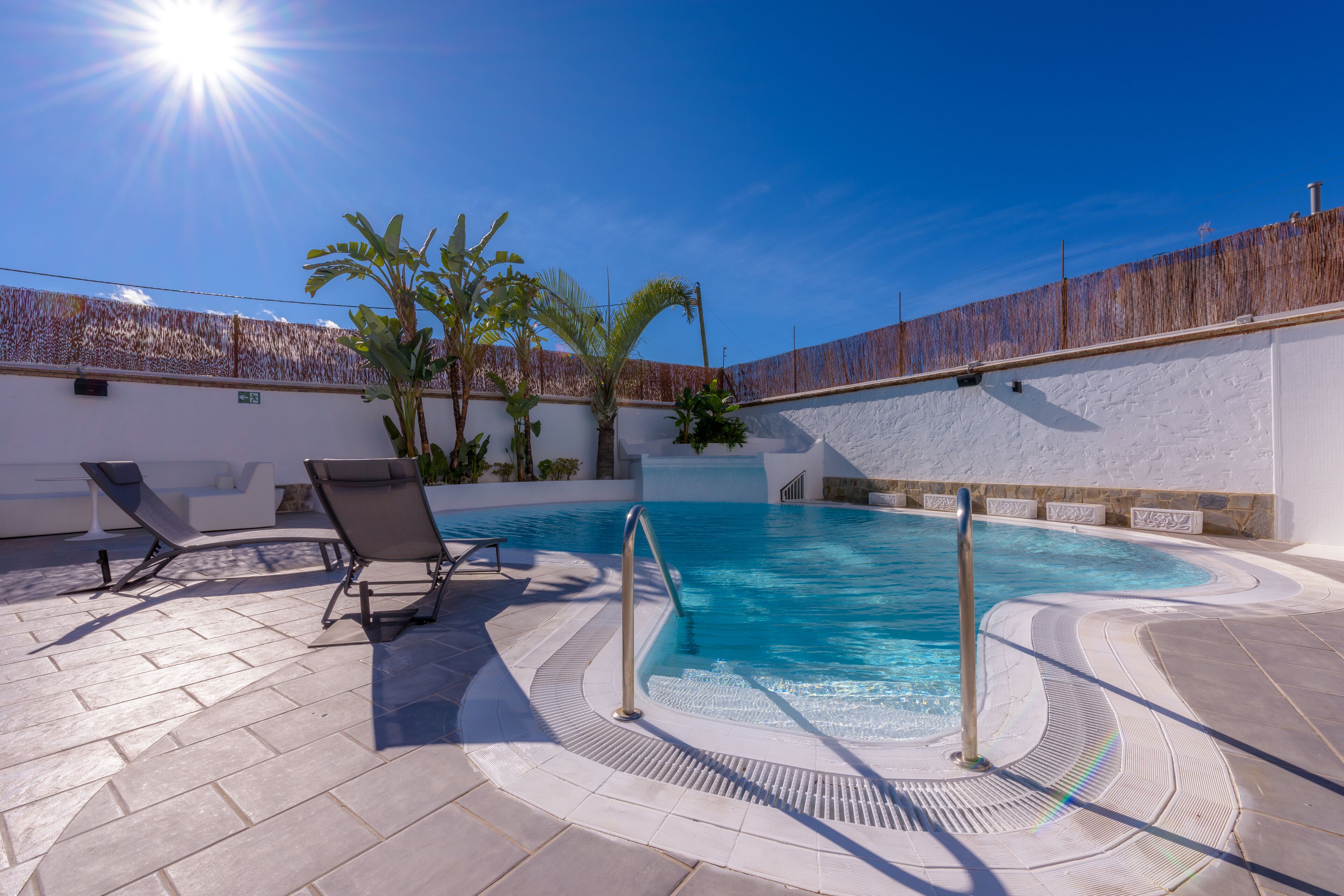 Un área de piscina con cielo azul despejado, dos sillas de descanso y una palmera.