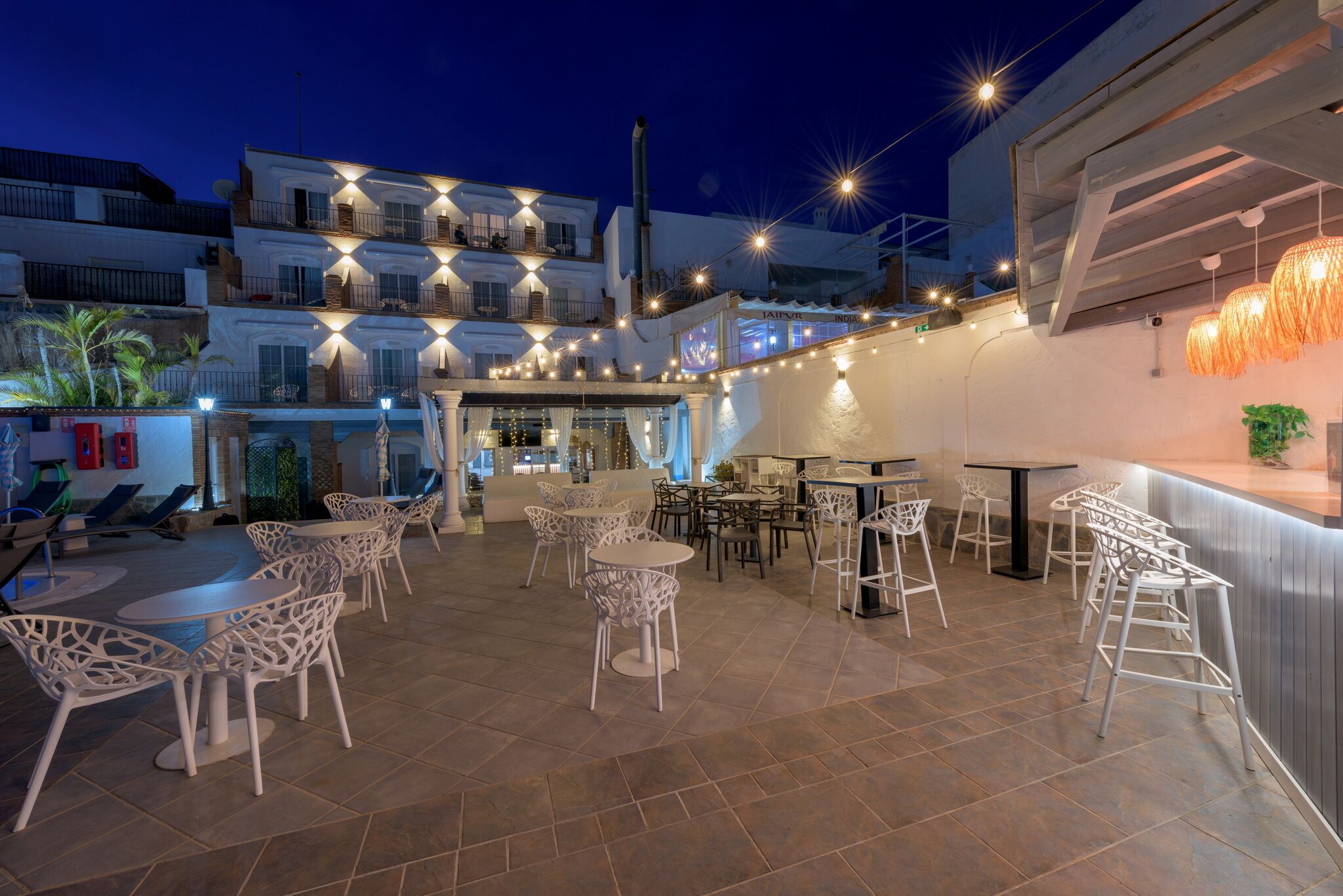 Un espace extérieur aménagé en terrasse, avec plusieurs coins salon, dont des chaises et des tables blanches, ainsi qu’un bar avec des tabourets.