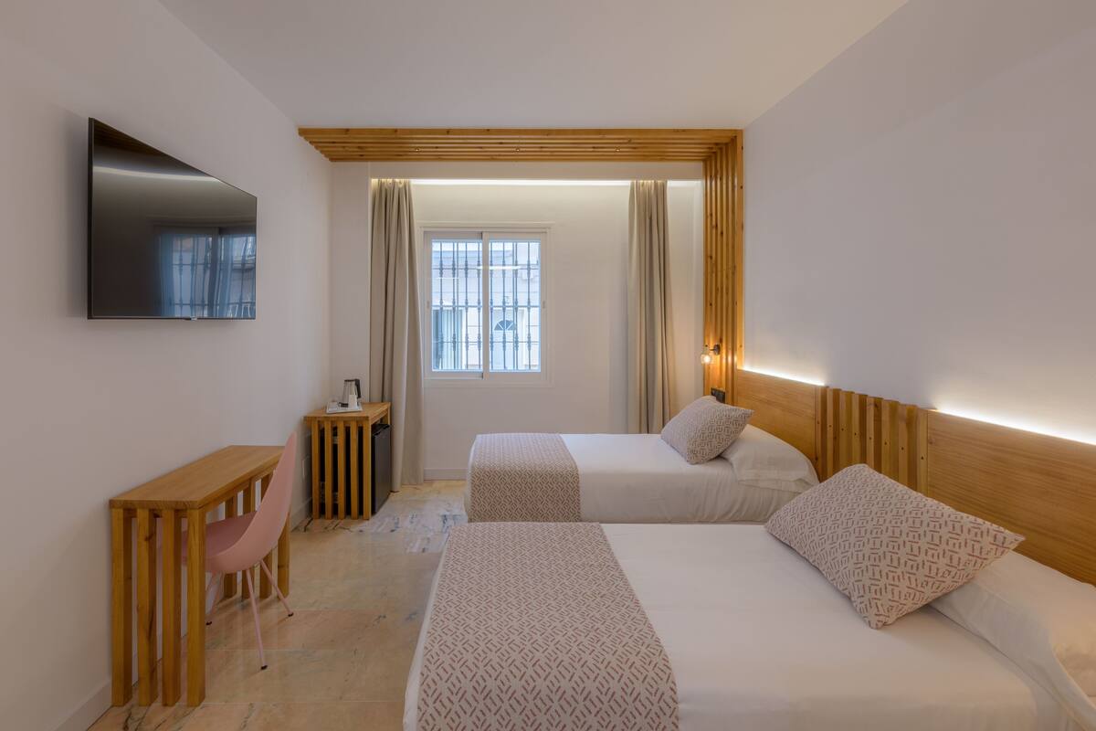 Une chambre d’hôtel avec deux lits, un téléviseur fixé au mur, une petite table en bois, une chaise et une fenêtre avec des rideaux.