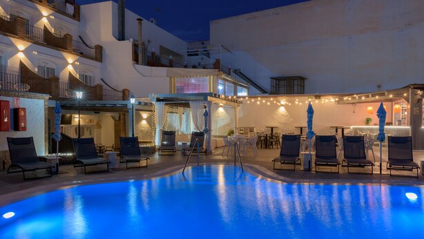 A swimming pool with lounge chairs and umbrellas, surrounded by buildings with balconies and string lights.